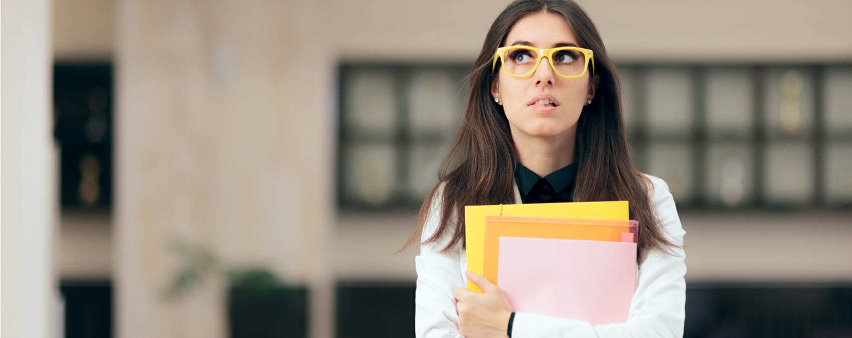A woman wearing glasses and holding a stack of papers bites her lip and looks like she is thinking intently