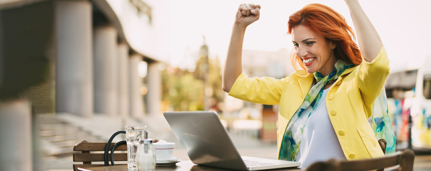 Woman smiles with her hands held above her head triumphantly while looking at a laptop