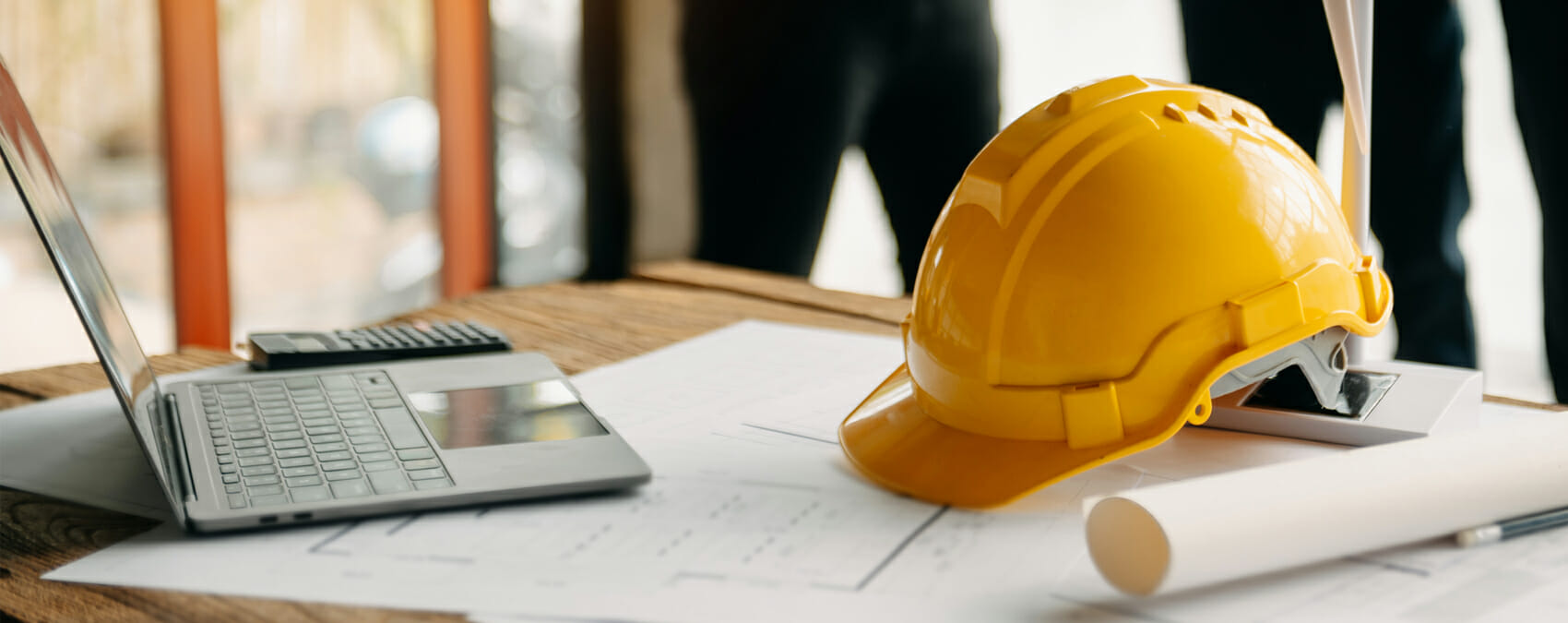 A hard hat sits on a desk next to a laptop and blueprints